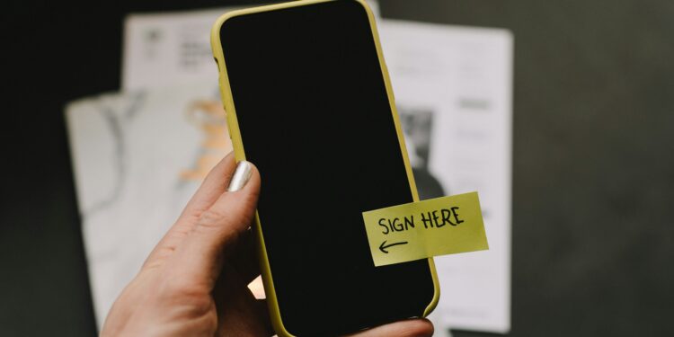 Ein Handy mit einem Post-it wird in die Kamera gehalten; auf dem Post-it steht “sign here”.